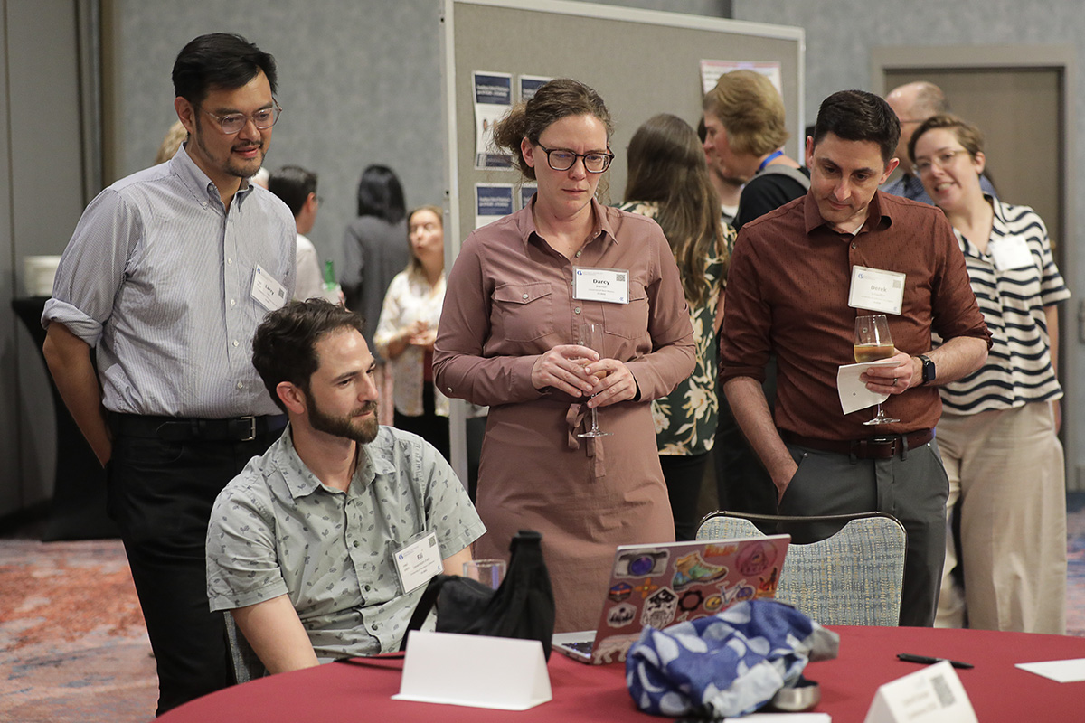 Three people standing and one seated huddled around a laptop on a table at the at the 2026 Cottrell Scholars Conference