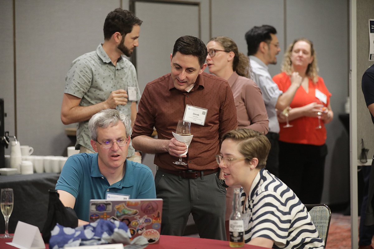 Two people seated and one standing huddled around a laptop on a table at the 2026 Cottrell Scholars Conference