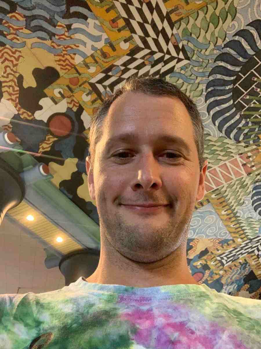 portrait of Jay Foley, associate professor professor of chemistry, wearing a tie-dyed t-shirt and looking down at the camera with a ceiling painted with artistic and graphical elements behind him
